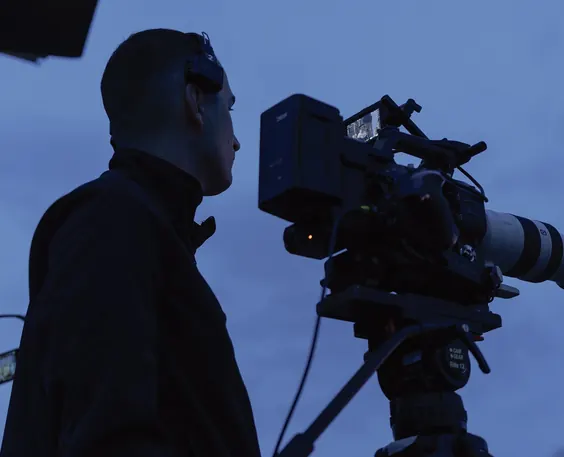 A cinematographer behind a film camera at dusk, in silhouette against a blue sky